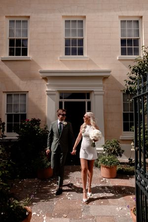 Bride in mini dress holding white bouquet walks with groom in green suit outside elegant Georgian building.