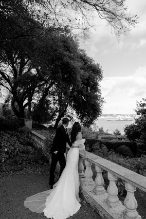 A couple stands on an ornate balcony overlooking a scenic waterfront view.