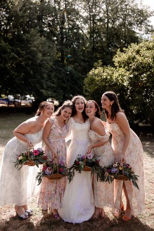 A bridal party in floral and white dresses laughs together while sitting outdoors in natural sunlight.
