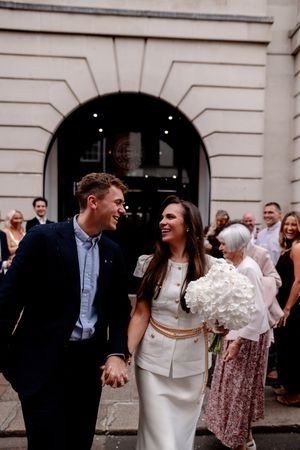 Smiling newlywed couple holding hands outside a grand building, bride in white suit holding hydrangea bouquet with guests behind.