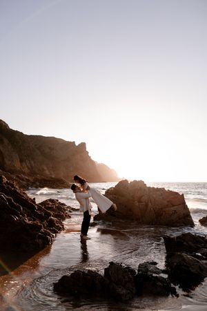 Couple embracing on rocky beach at sunset with ocean waves and cliffs in background.