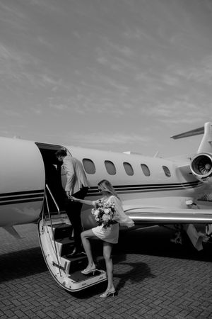 Black and white photo of a bride holding flowers boarding a private jet with a companion on the stairs.
