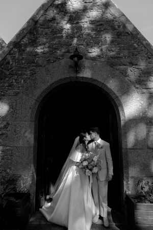 Black and white photo of bride and groom kissing in arched stone church doorway, bride holding floral bouquet.