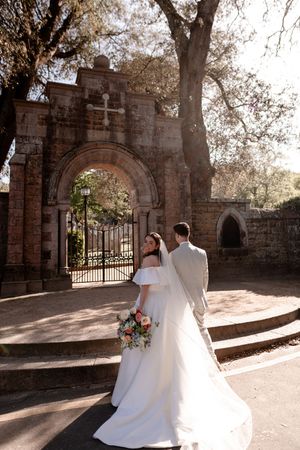 Bride and groom walk toward ornate brick archway gate with cross, bride holding colorful bouquet in white gown.