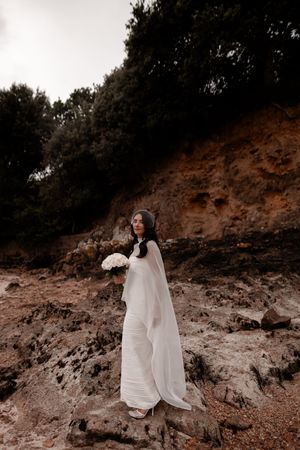Bride in white gown and veil holding bouquet on rocky coastal cliff with dark trees backdrop.