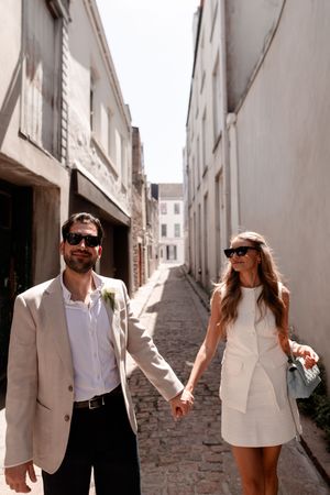 Stylish couple in sunglasses holding hands walking down a charming cobblestone alley in matching white and beige wedding attire.
