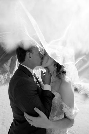 Black and white photo of a bride and groom sharing a romantic kiss under a flowing wedding veil.