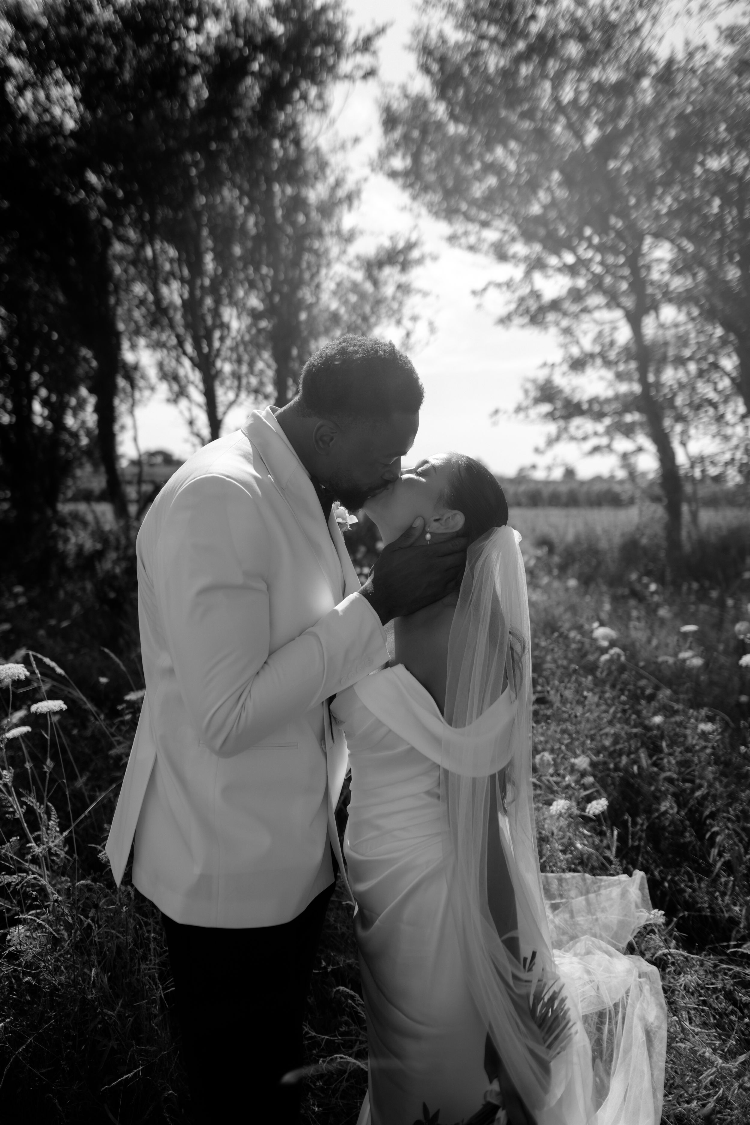 Black and white photo of a couple sharing a romantic kiss outdoors, bride in white gown and veil, groom in white shirt.