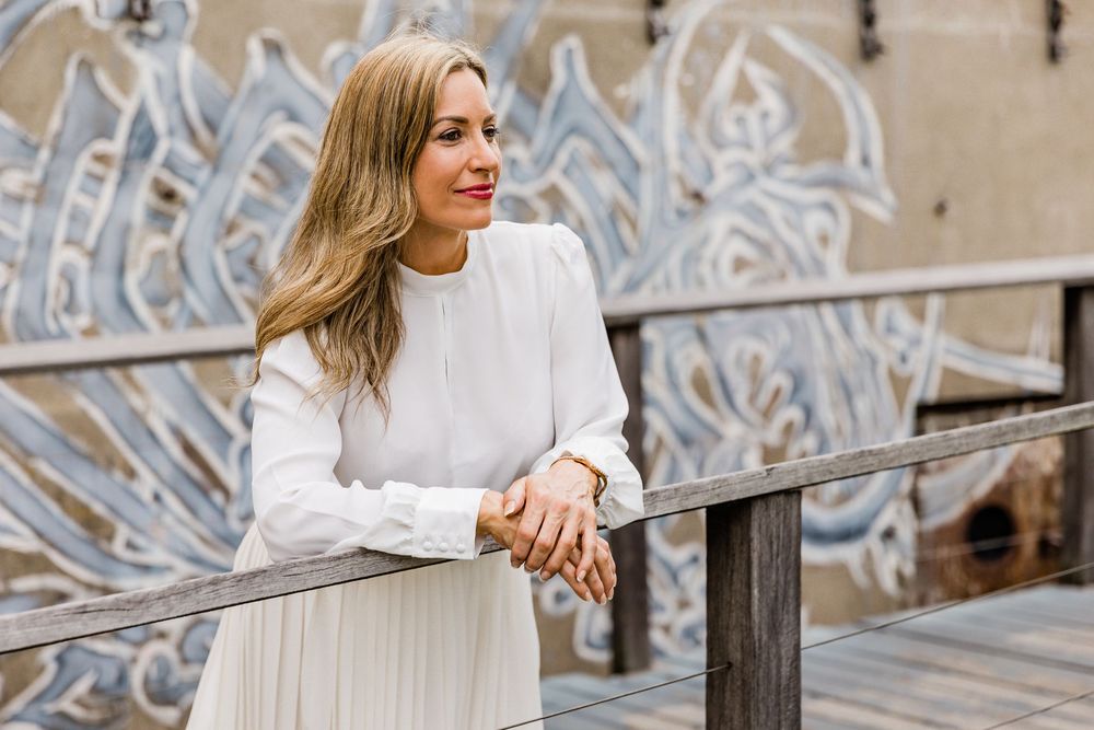 A person in a white sweater stands against a textured wall with decorative architectural details in the background.