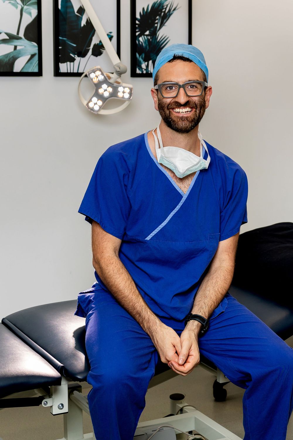 Healthcare professional in blue scrubs and surgical mask sits on examination table in medical office.