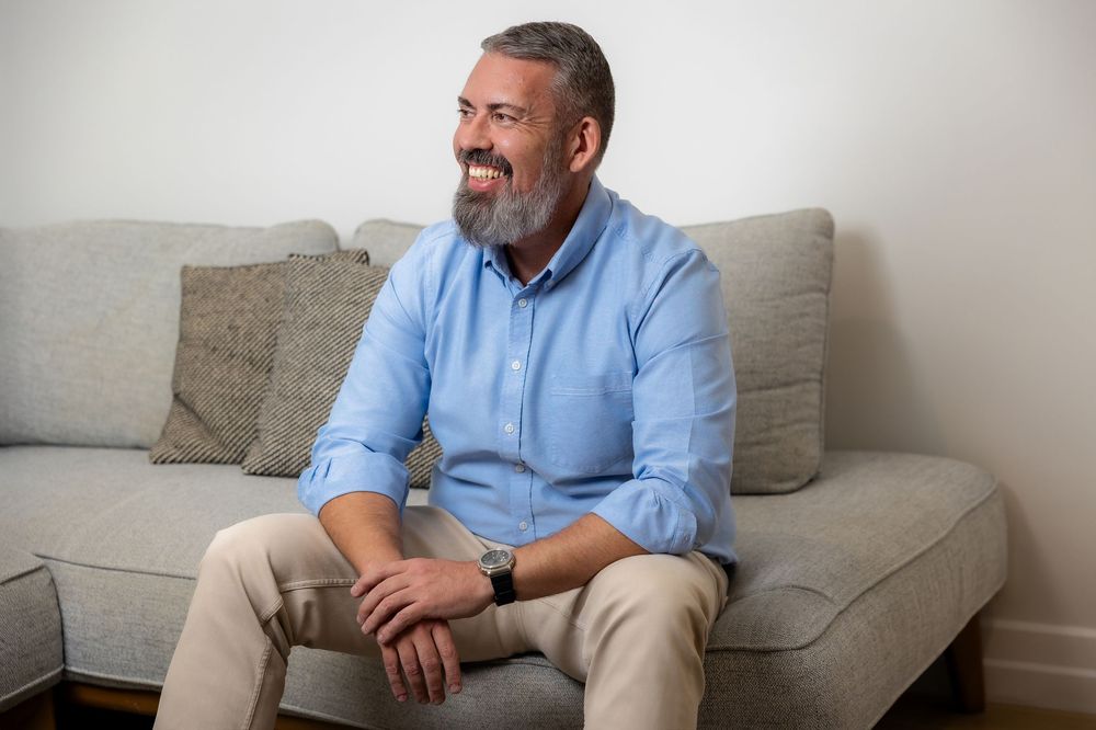 Middle-aged man with gray beard sitting on gray sofa, smiling, wearing blue shirt and khaki pants.