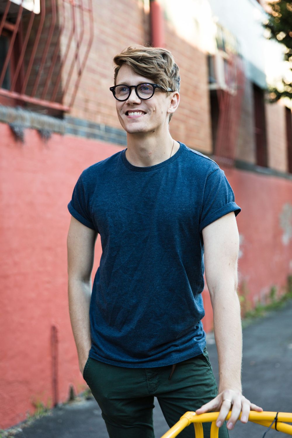 Person wearing navy blue t-shirt and dark pants smiles while sitting against coral-colored brick wall background.
