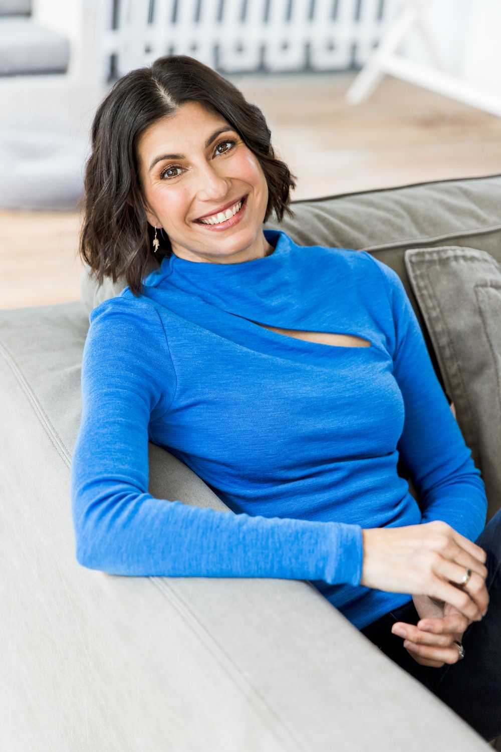 Smiling person wearing a bright blue long sleeve top with cutout detail relaxes on a gray couch.