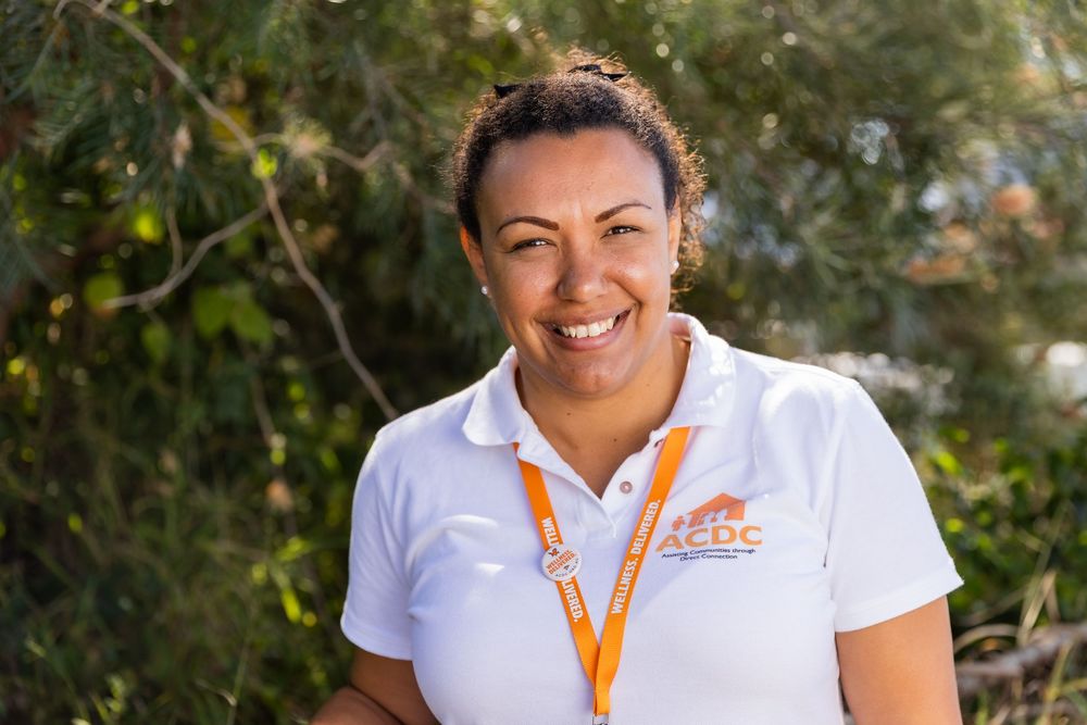 A person in a white polo shirt with orange accents smiles while standing outdoors with greenery in the background.