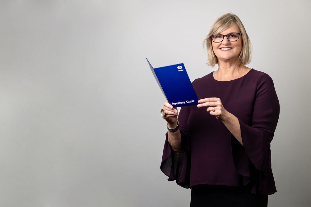 A person wearing glasses and a purple sweater holds a blue folder while standing against a light gray background.