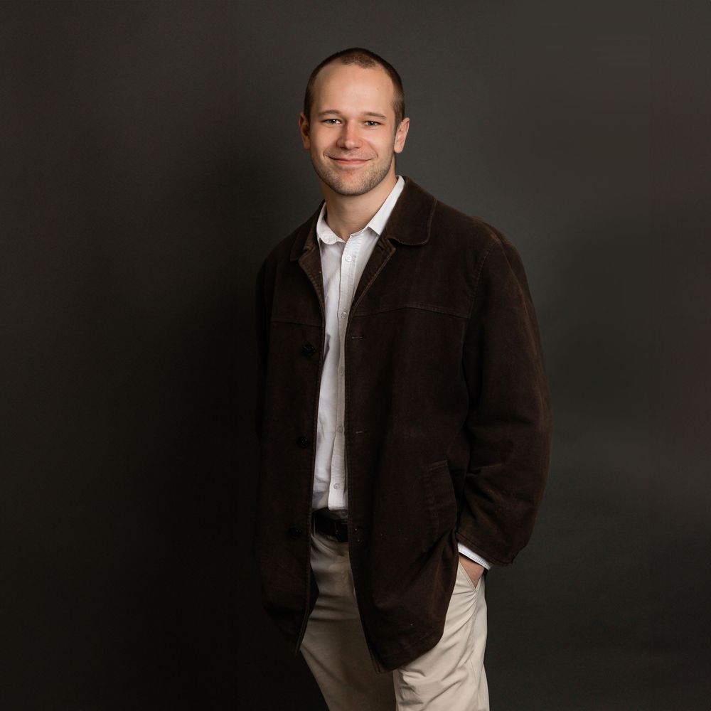 Professional studio portrait of a person in a brown jacket and white shirt against a dark background.