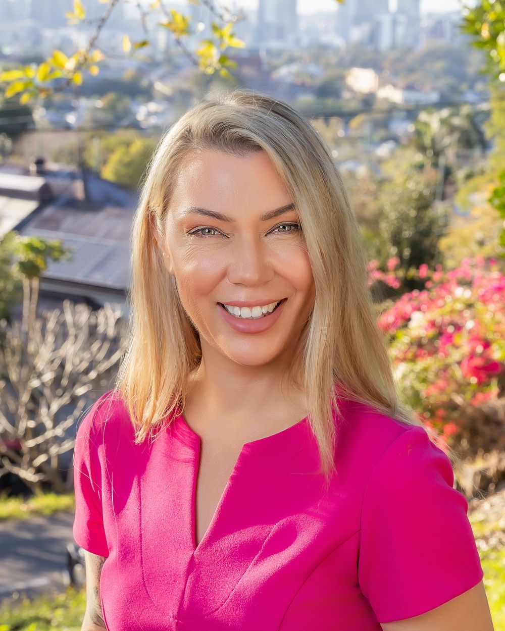 A person wearing a bright pink top smiles outdoors with blooming flowers and city buildings in the background.