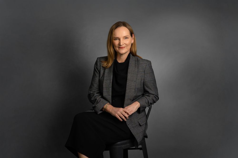 Professional portrait of someone wearing a dark blazer against a grey background with dramatic lighting.