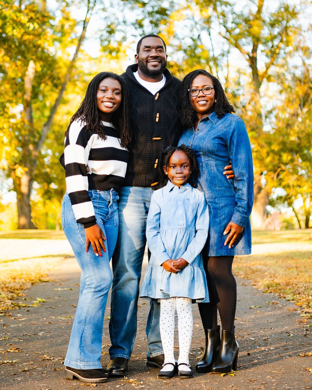 Family of four poses together on autumn path with golden leaves in background.