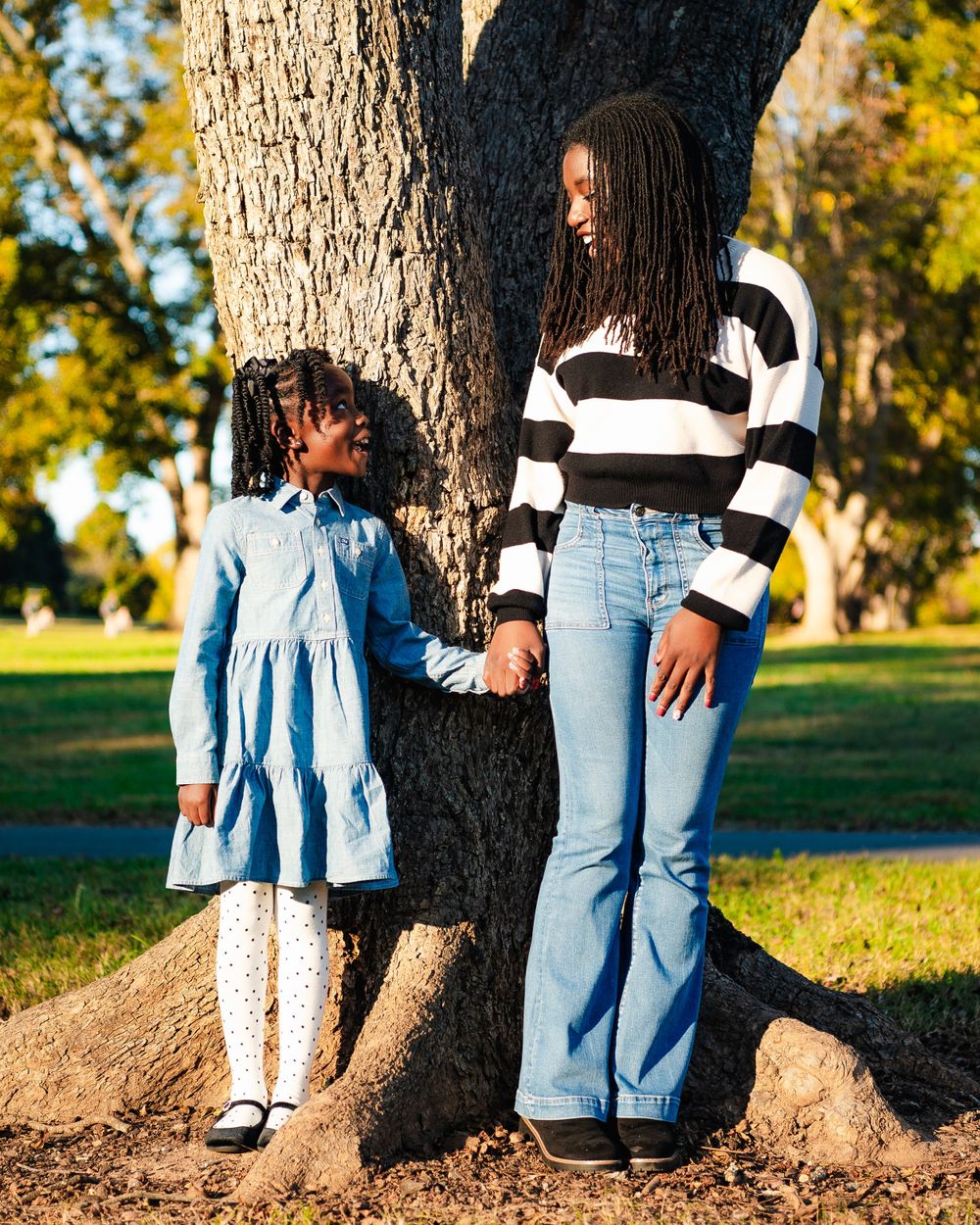 Parent and child holding hands while standing against large tree trunk in park.