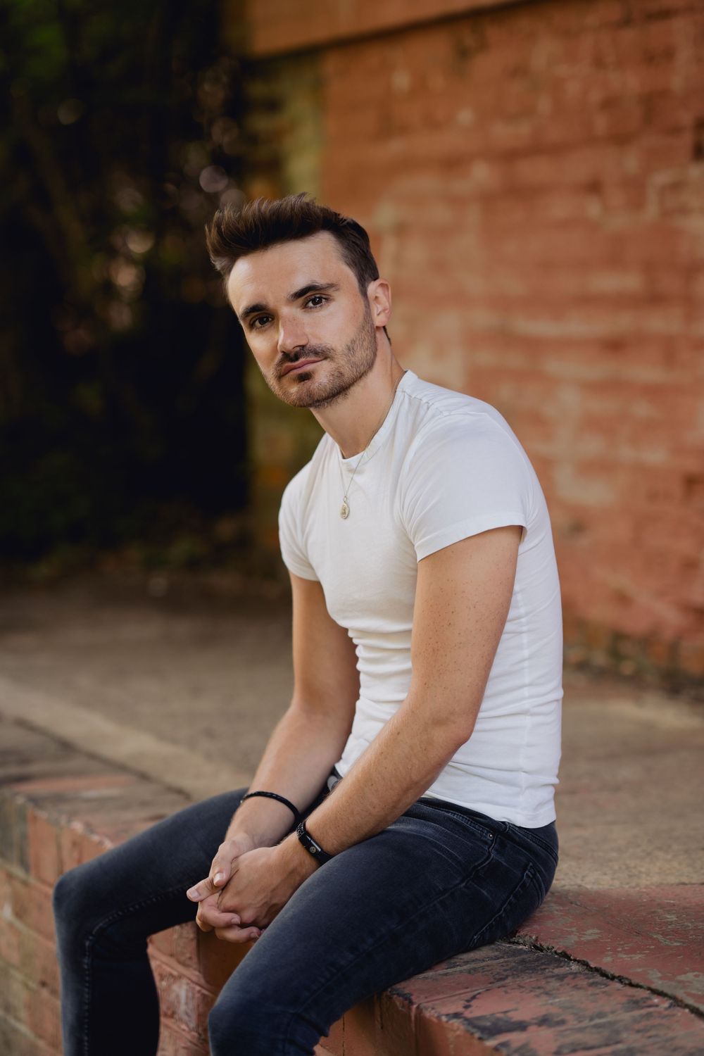 Person in white t-shirt and dark jeans sits on brick steps in outdoor setting at dusk.