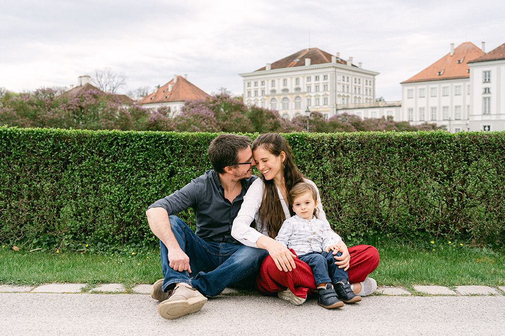 A family sits together on grass near a beautiful European-style building with hedges.