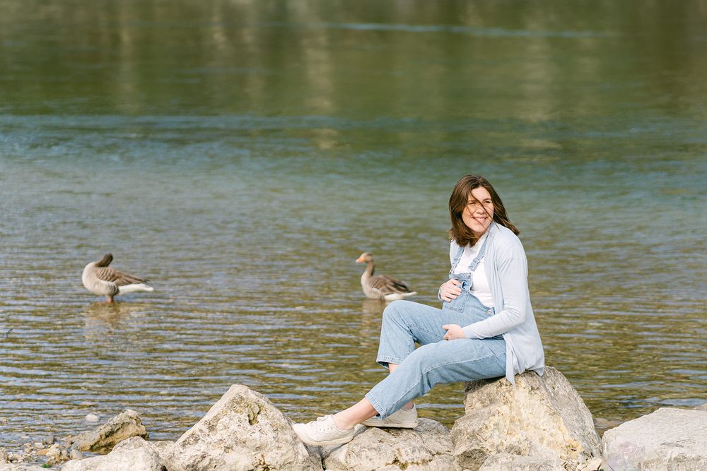 Person in white shirt sitting on rocks by water with ducks swimming nearby.