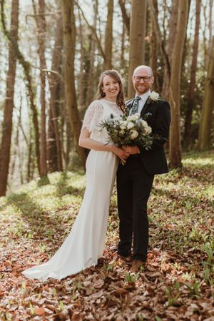 Newlyweds pose in a wooded forest setting holding a white floral bouquet.