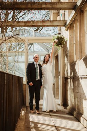 A couple poses in formal wedding attire under a wooden pergola with natural light streaming through.
