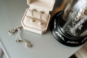 Wedding jewelry and accessories including a beige ring box and delicate silver necklace on a gray surface.