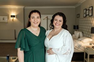 Two women in elegant dresses, one in emerald green and one in white, smile warmly in an indoor setting.