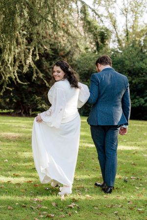 A couple in wedding attire walks away from camera across a green lawn with trees in the background.