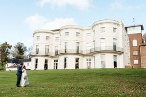 A couple poses on the lawn of a grand white Georgian-style mansion during their wedding day.