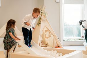 Children play in a row of white teepee-style tents set up in a bright indoor playroom.