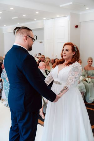 Bride and groom hold hands during wedding ceremony, bride wearing lace dress with guests seated behind.