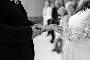 Black and white wedding photo of couple exchanging rings, bride with lace sleeve and tattoo holding groom's hands.