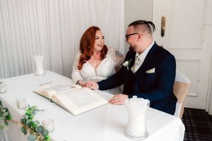 Bride and groom smiling at each other while signing wedding register at white table with greenery decor.