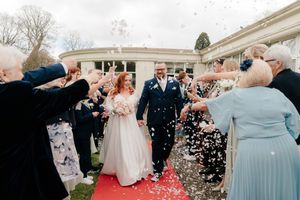 Bride and groom walk through confetti shower on red carpet surrounded by cheering wedding guests outside elegant venue.