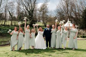 Bride and groom pose with bridesmaids in sage green gowns holding white bouquets aloft in a garden setting.