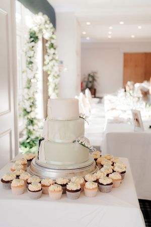 Elegant three-tier white wedding cake surrounded by cupcakes on a silver stand in a bright decorated reception venue.