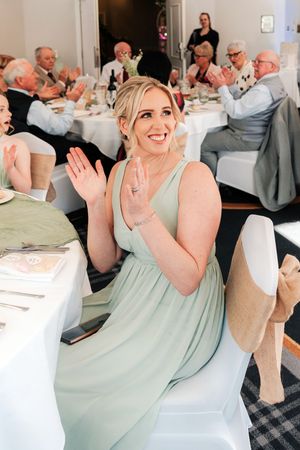Smiling bridesmaid in sage green dress clapping at wedding reception dinner.