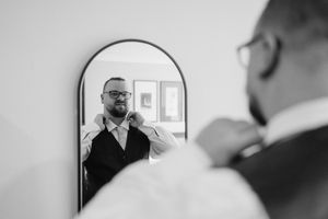 Groom adjusting his tie in an arched mirror, black and white wedding preparation photo.