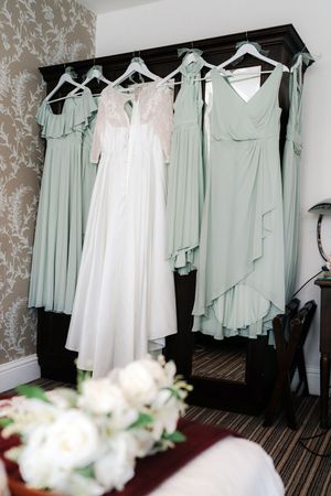 White wedding gown and sage green bridesmaid dresses hanging on a dark wardrobe in an elegant bridal suite.