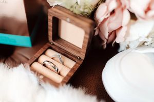 Two silver wedding rings displayed in an open wooden ring box, surrounded by pink roses and soft white fabric.