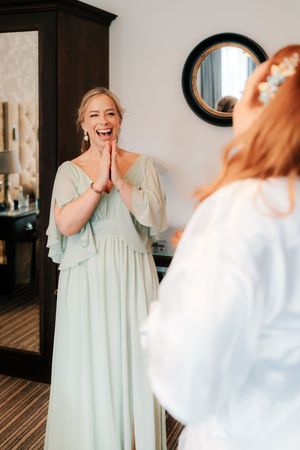 Bridesmaid in mint green gown reacts with joy seeing bride in white dress during wedding preparations indoors.