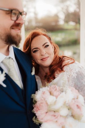 Smiling redhead bride leans on groom's shoulder holding pink peony bouquet at outdoor wedding.
