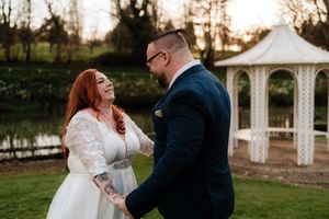 A joyful bride in lace wedding dress laughs with her groom in navy suit beside a white gazebo in a garden.