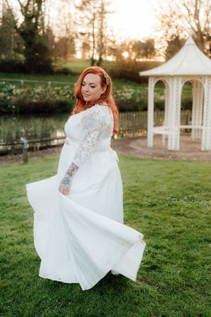 Red-haired bride in white lace wedding dress twirling on green lawn near gazebo at golden hour.