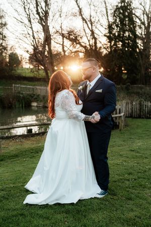 Bride and groom share romantic sunset moment by pond, bride in lace gown, groom in navy suit.