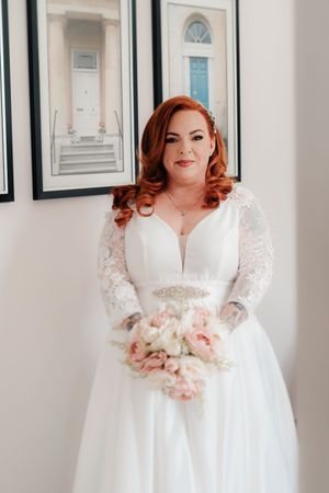 Bride in white lace long-sleeve wedding gown holding pink bouquet, posing indoors.